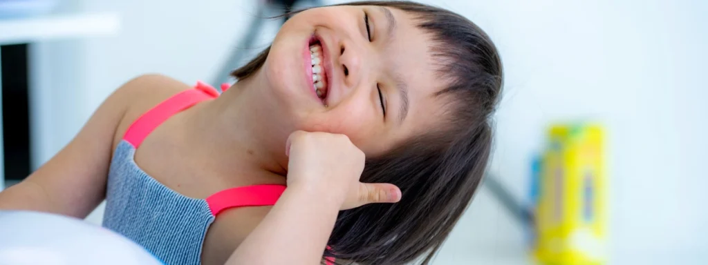 A young girl with Down syndrome wearing a striped dress with pink straps smiles brightly while tilting her head and making a thumbs-up gesture. The background is blurred with bright indoor lighting.