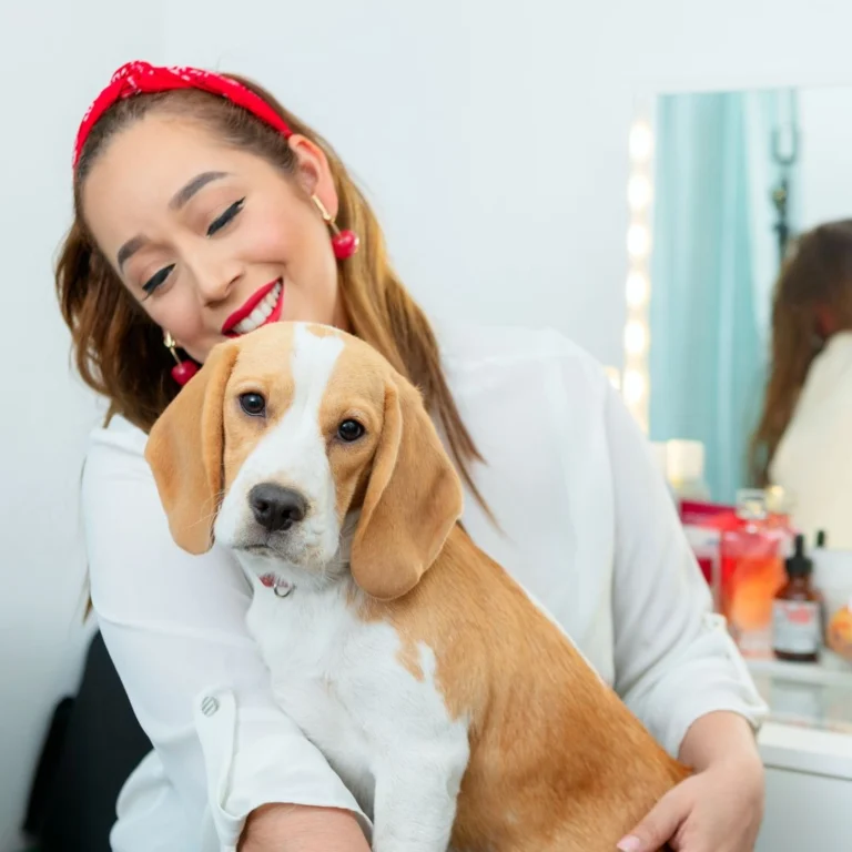 A smiling woman wearing a red headband and white blouse hugging a beagle puppy in a well-lit room with a vanity mirror in the background.