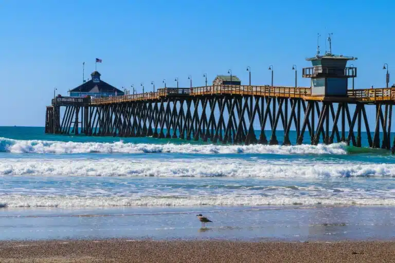 Imperial Beach shoreline with sandy beach, gentle ocean waves, and a long wooden pier extending into the Pacific under a bright sky.