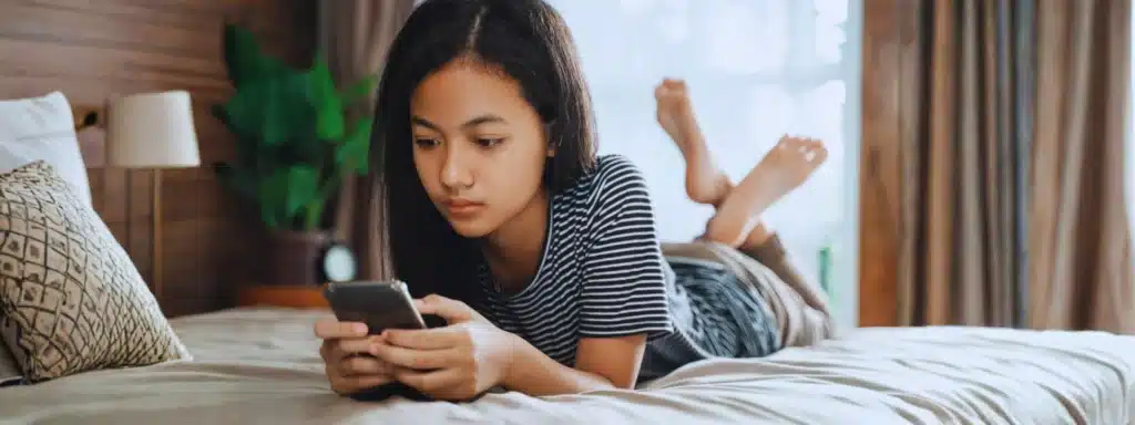 Teen girl lying on bed looking at smartphone, reflecting emotional and behavioral health challenges.