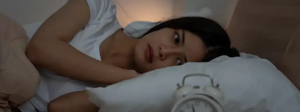 Woman lying awake in bed at night, staring at an alarm clock, illustrating anxiety, insomnia, and fear of falling asleep