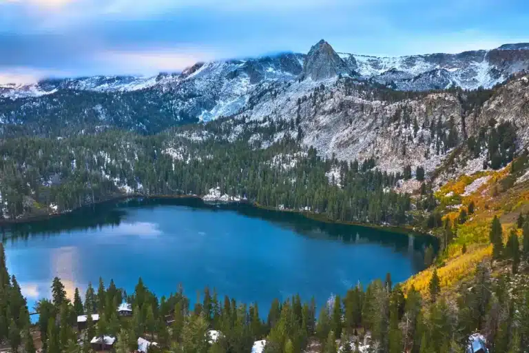 Scenic view of an Alpine, California mountain lake surrounded by pine forests and rugged rocky peaks under a blue sky.