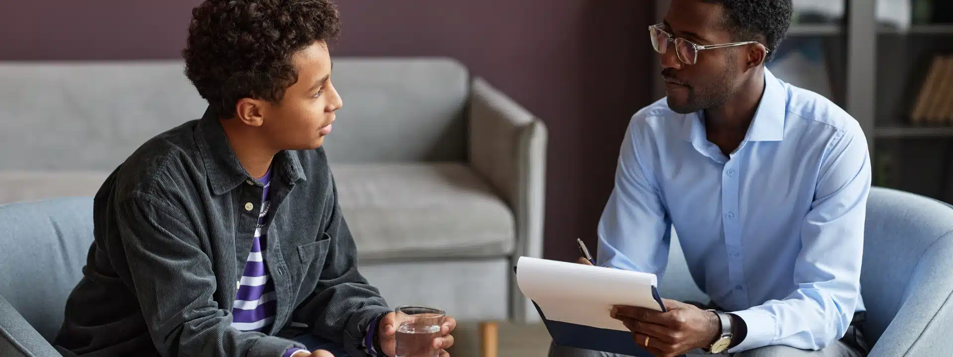 A young person sits in a chair holding a glass of water while speaking with a counselor or therapist who is taking notes during a session in a comfortable office setting.