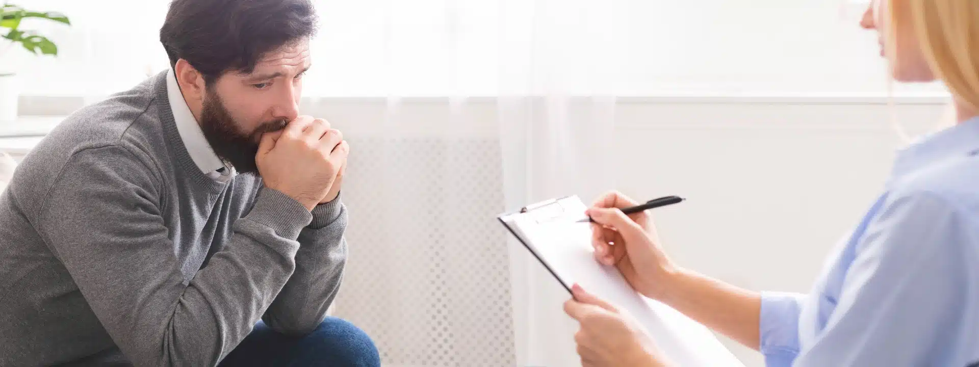 A man sits in front of a woman holding a clipboard, engaged in a discussion or interview setting.
