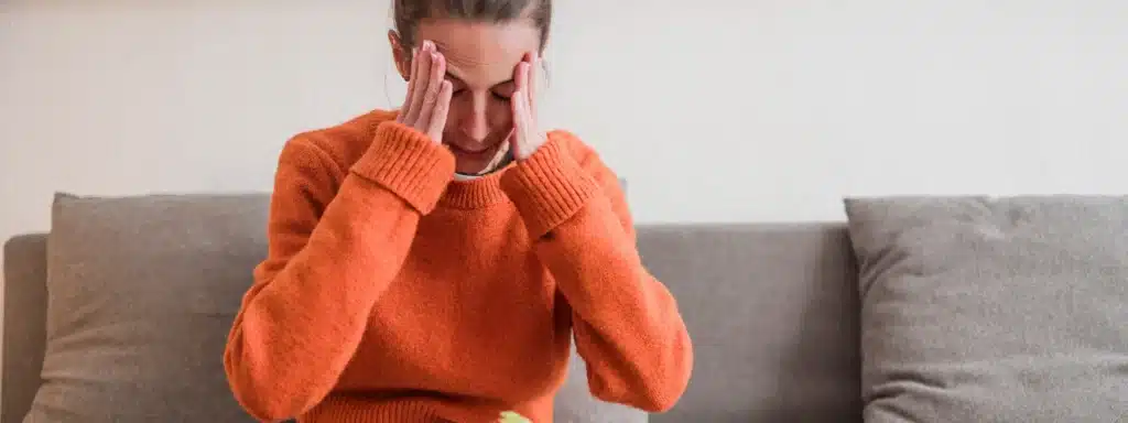 A woman sits on a couch, resting her hands on her face, conveying a sense of contemplation or distress.