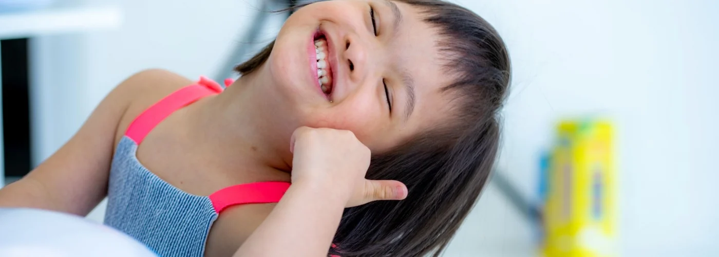 A young girl with Down syndrome wearing a striped dress with pink straps smiles brightly while tilting her head and making a thumbs-up gesture. The background is blurred with bright indoor lighting.