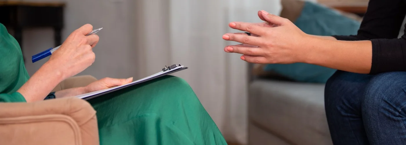 Close-up of a therapy session showing a seated therapist holding a clipboard and pen, while a patient gestures with their hands, emphasizing a conversational and supportive environment.