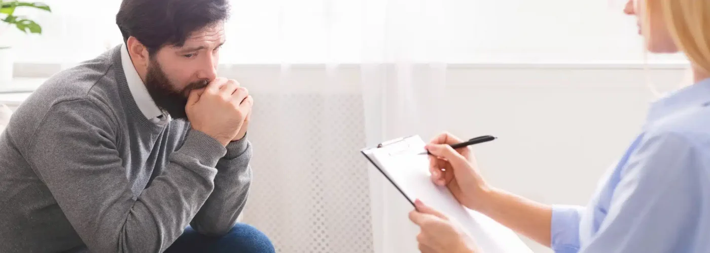 A man sits in front of a woman holding a clipboard, engaged in a discussion or interview setting.